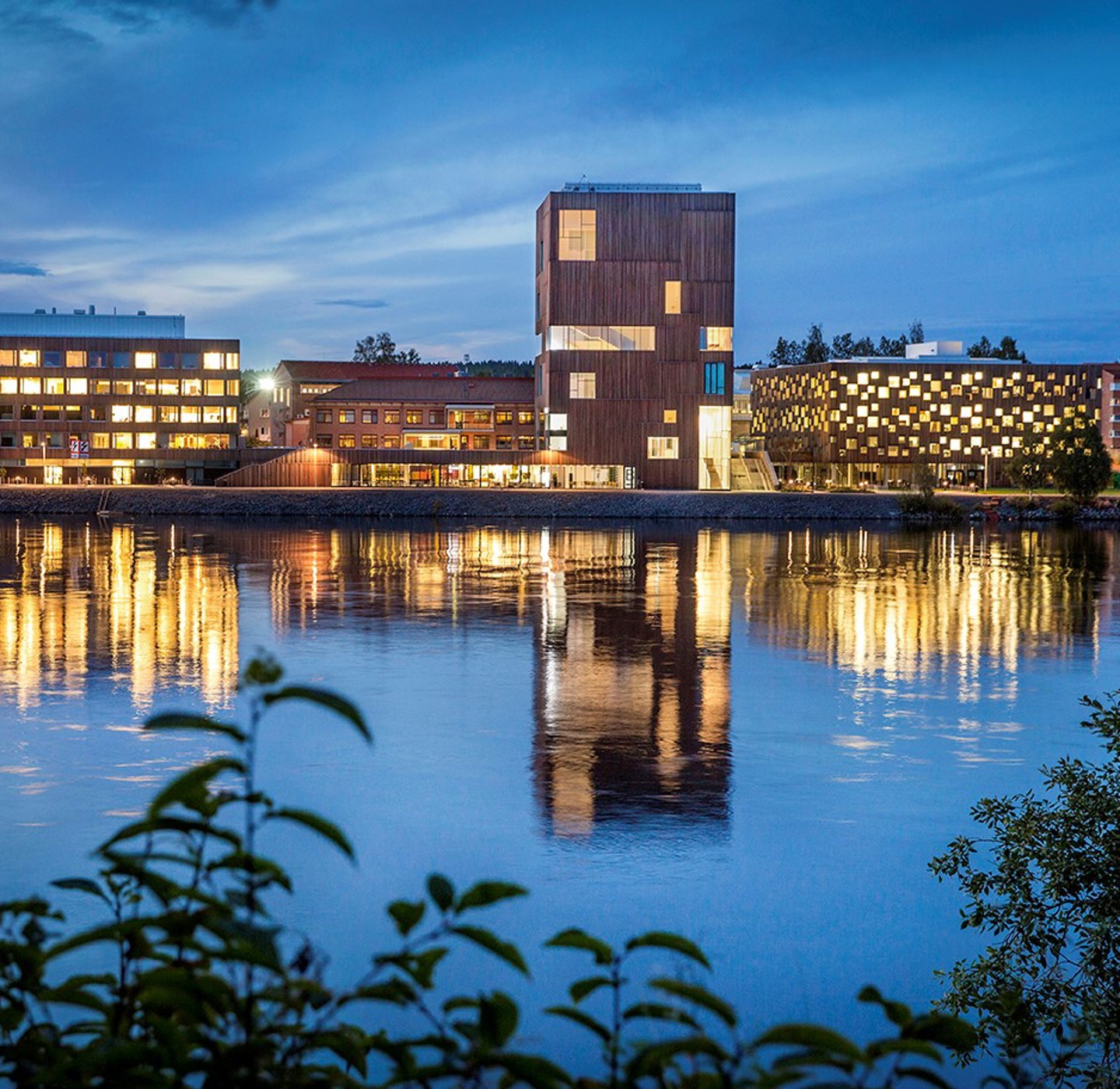 An evening photo of arty buildings. Lights from the windows are reflected on the water in the foreground. 