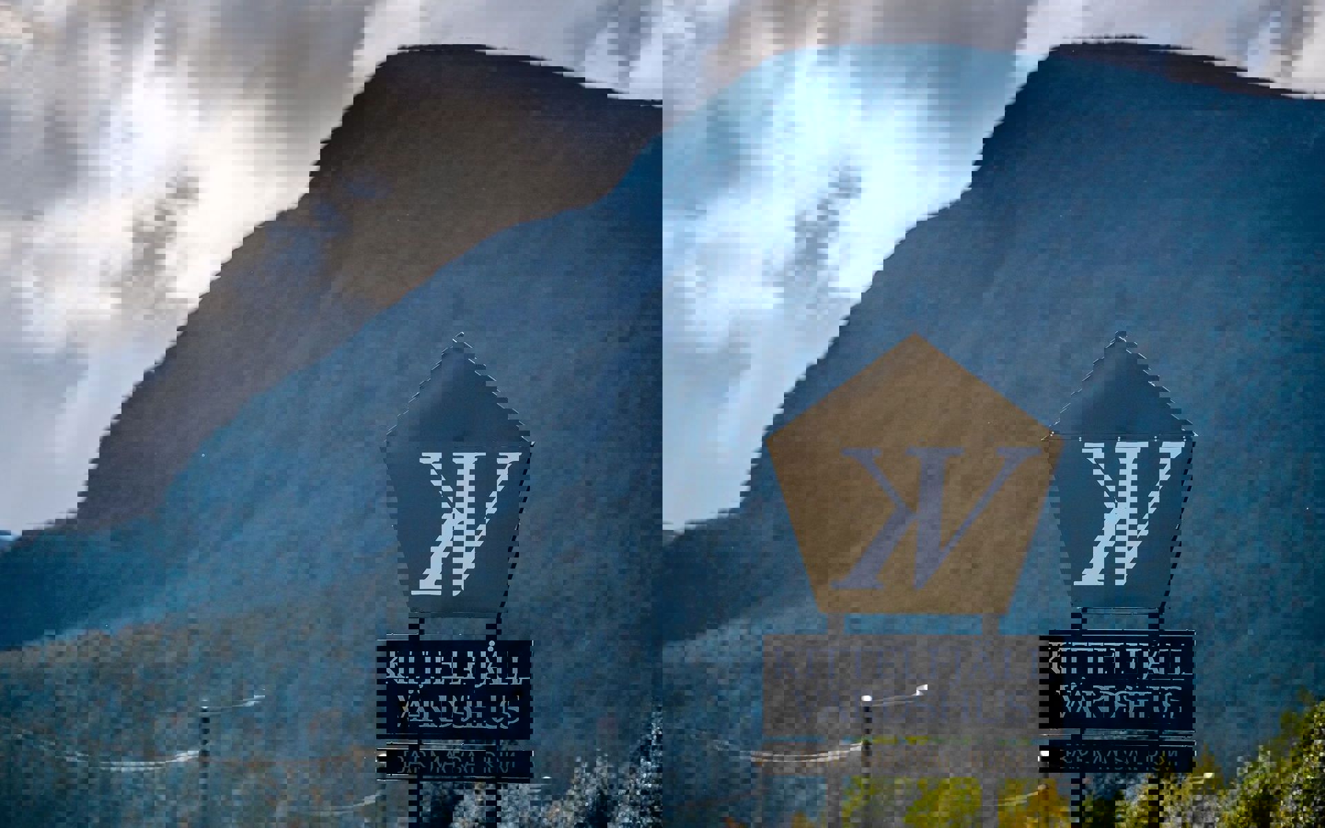 A restaurant sign in the foreground with a large green maountain in the background and a cloudy sky. 