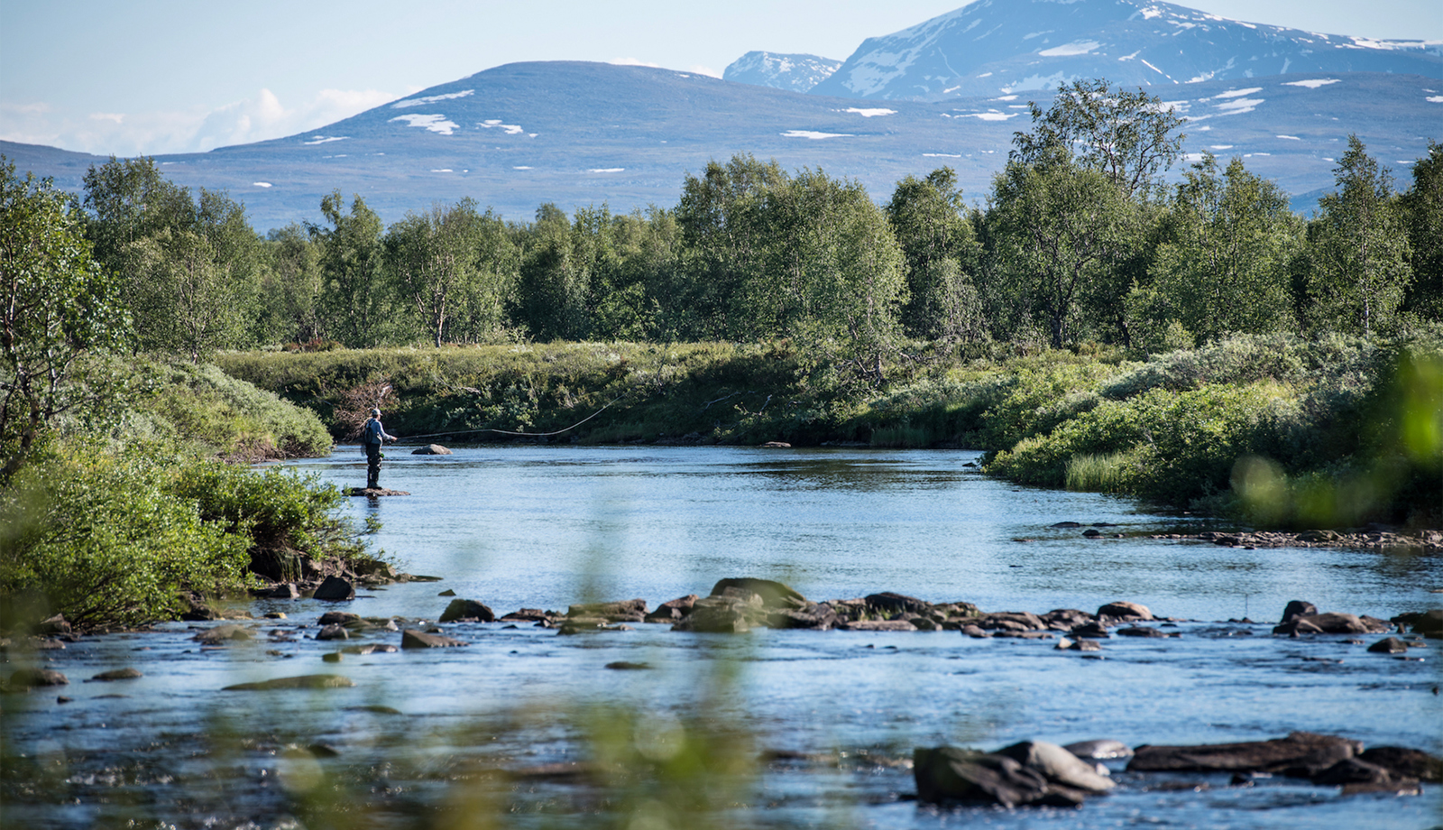 A person fishing in the river with mountains in the background. 