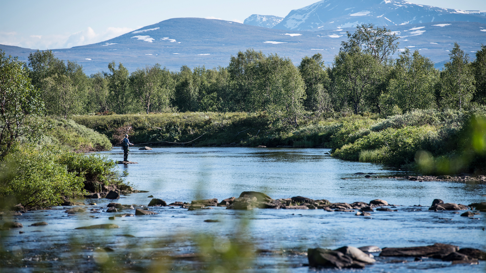 A person fishing in the river with mountains in the background.