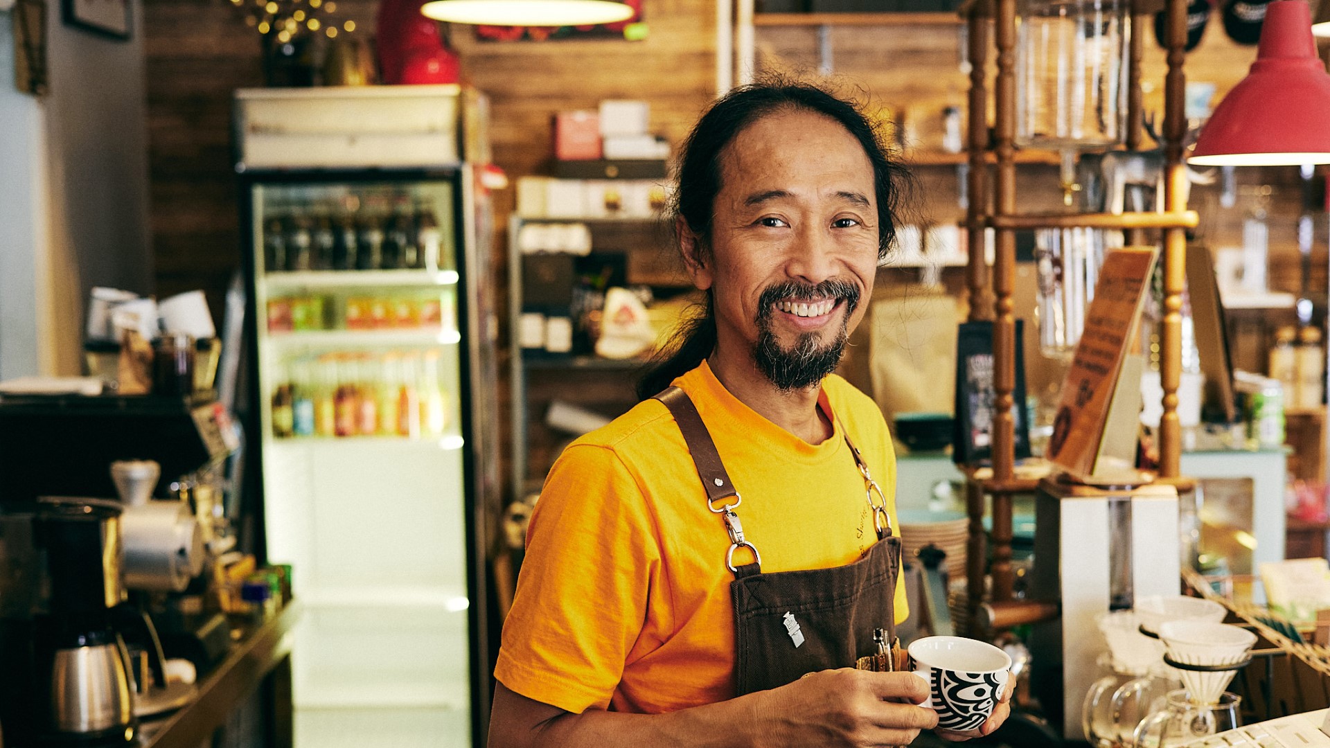 Budha's Coffee Roastery Foto: Per Jonsson @shotbyper A man in a yellow sweater holding a coffee cup, smiling towards the camera.