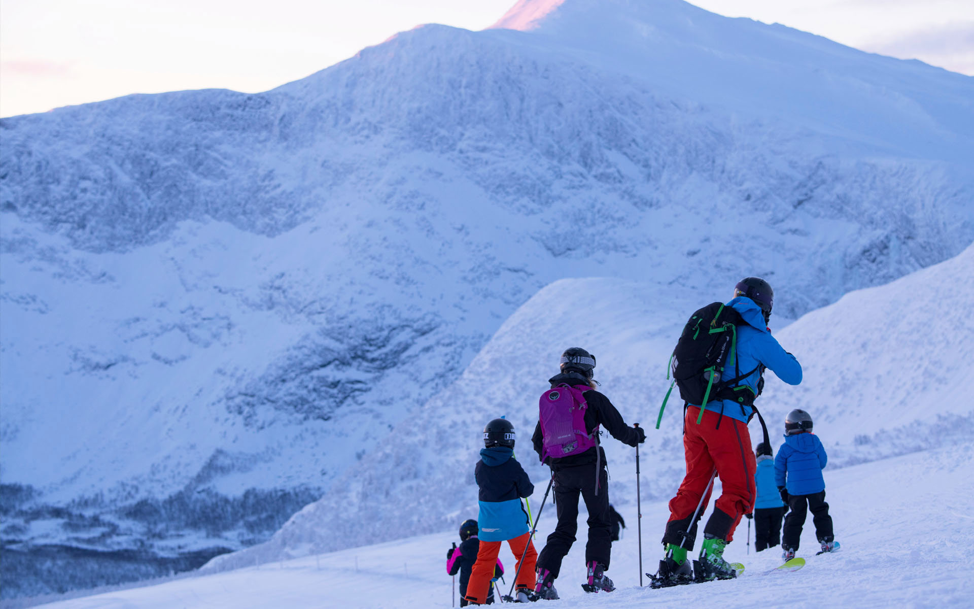 A skiing family standing with their backs towards the camera, a large steep mountain in the background. 